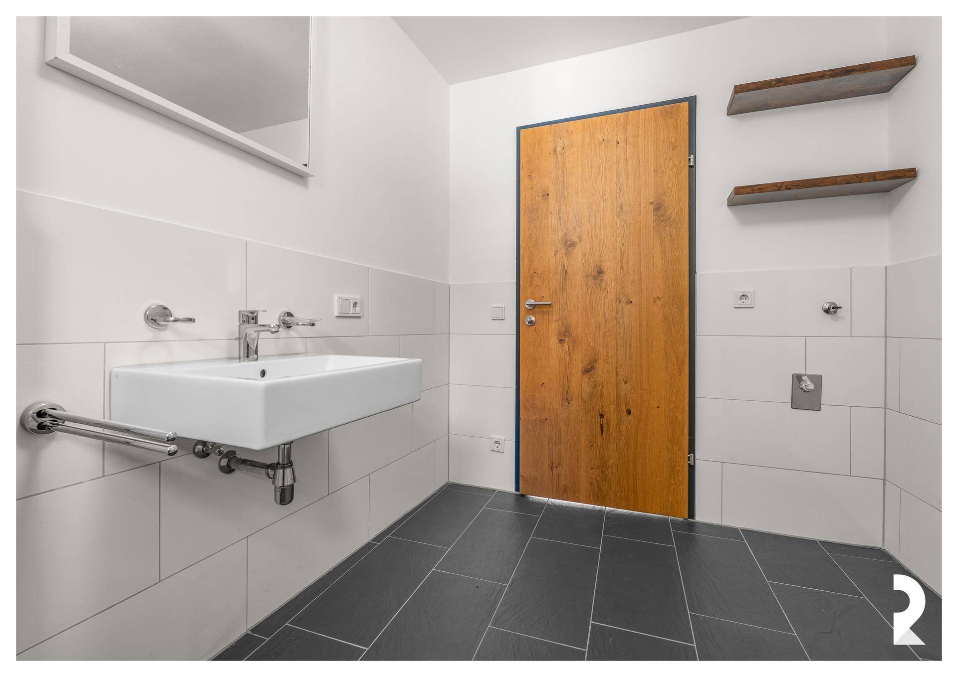 Modernes Badezimmer mit Waschbecken, Spiegel, Holztür und Haltegriff für zusätzliche Sicherheit.