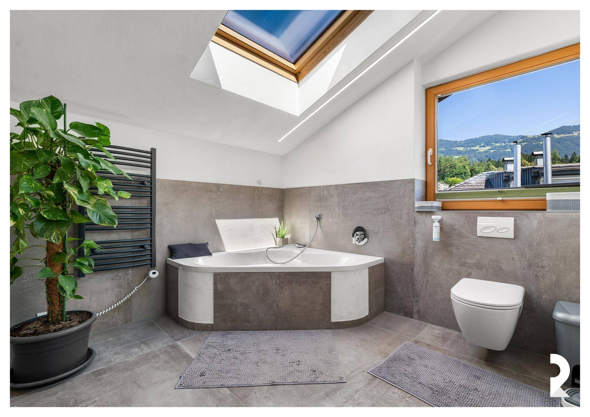 Modernes Badezimmer mit Eckbadewanne, Wand-WC, Dachfenster und Ausblick auf die Berge.