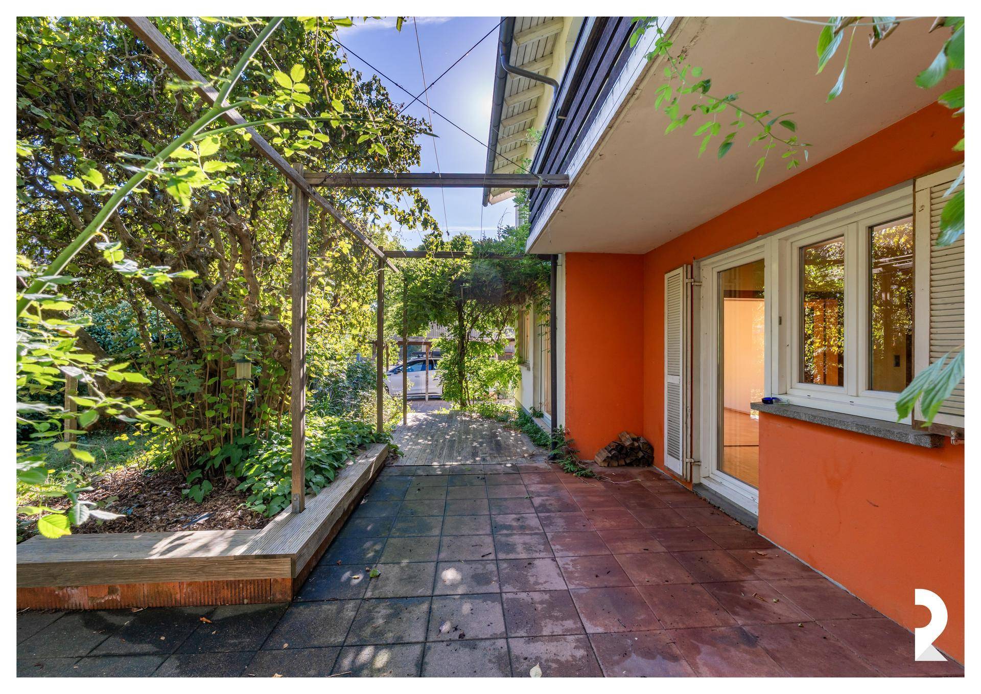 Gepflasterte Terrasse mit Pergola und üppigem Grün neben einem Haus mit oranger Fassade.