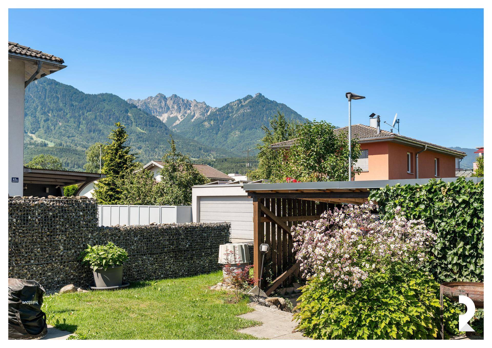 Gepflegter Garten mit Schuppen, Gabionenmauer und weitem Blick auf die Berge.