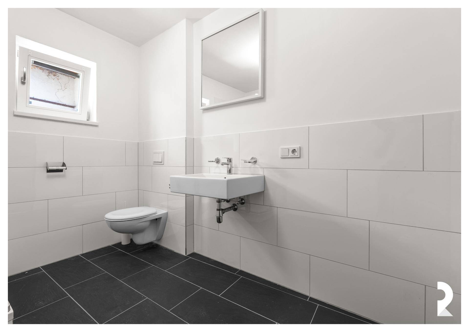 Helles WC mit Waschbecken, Spiegel, Fenster und dunklen Bodenfliesen, ideal für Gäste.