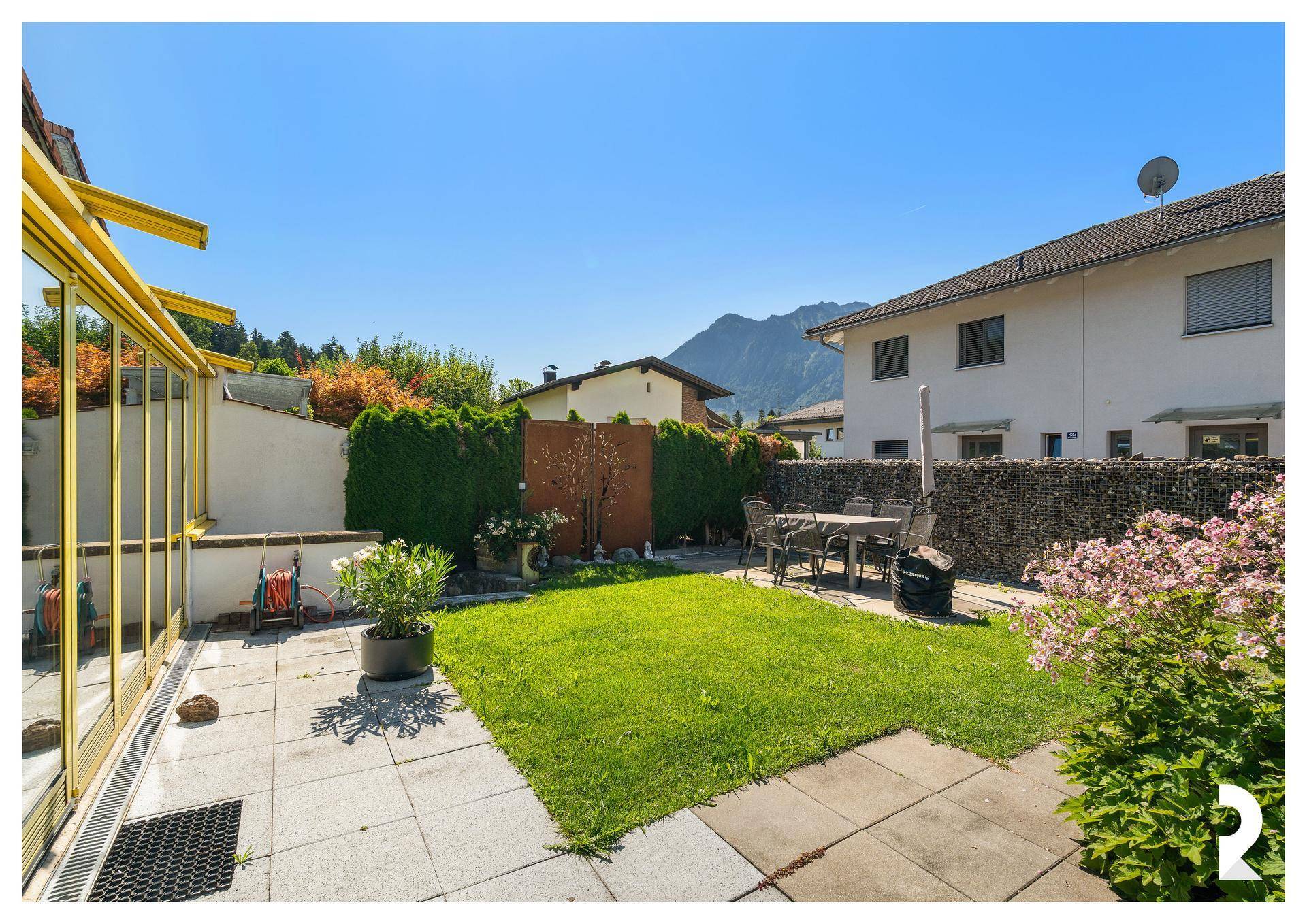 Sonniger Garten mit Rasenfläche, gepflasterter Terrasse und angrenzendem Wintergarten.