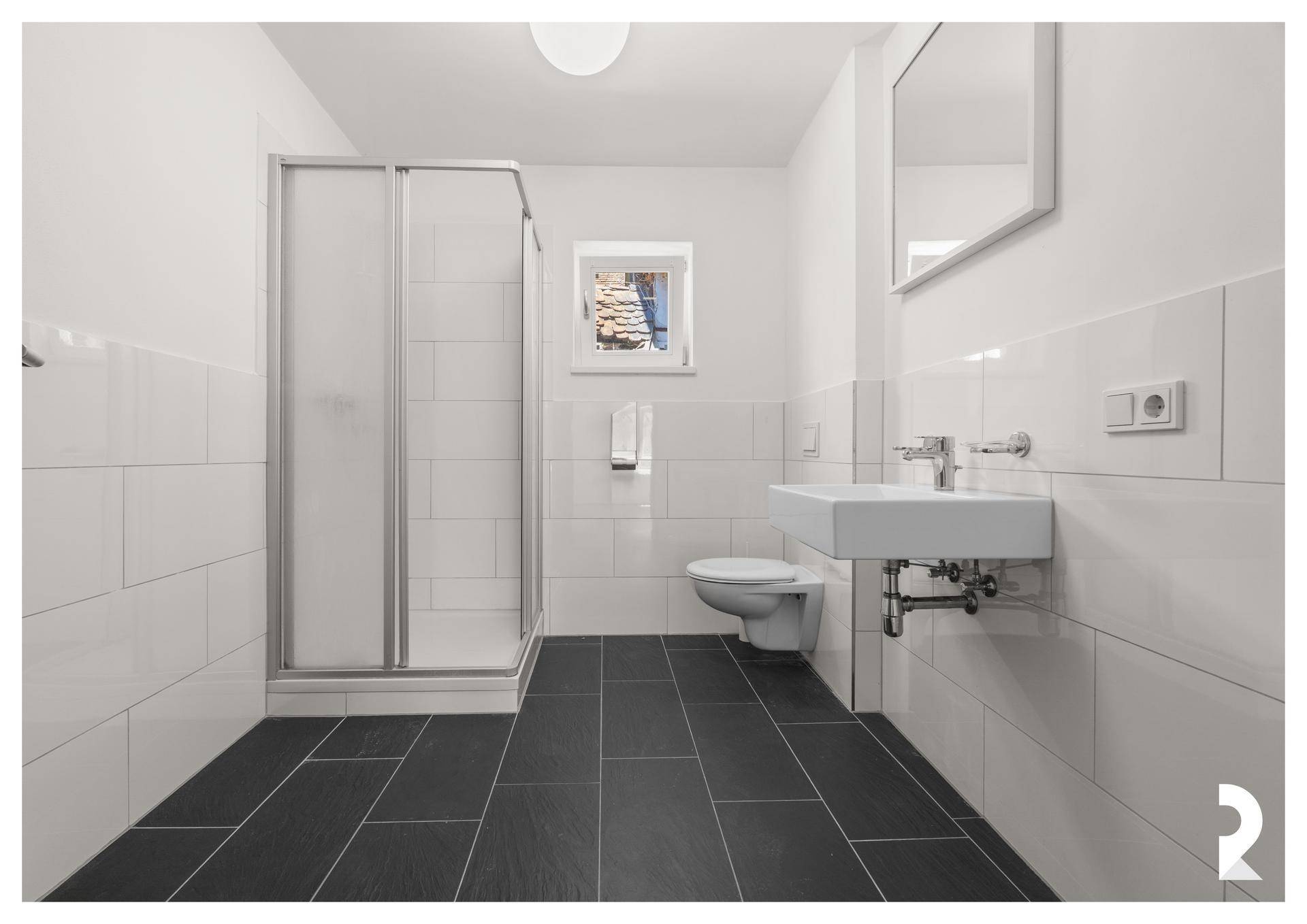 Modernes Badezimmer mit Dusche, Waschbecken, WC, Fenster und dunklen Bodenfliesen.