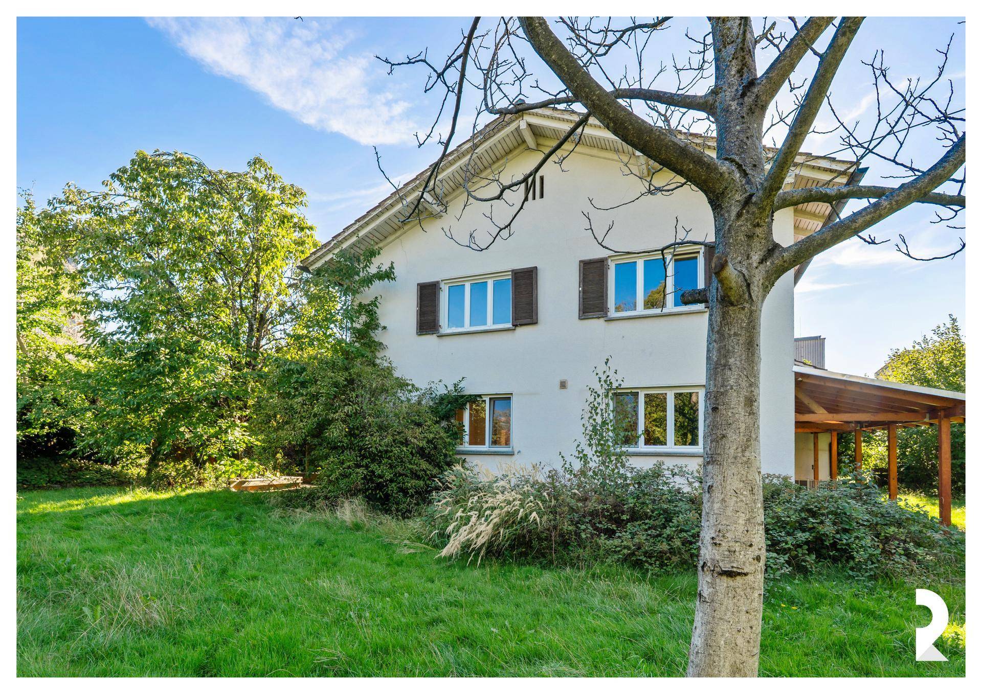 Weißes Einfamilienhaus mit braunen Fensterläden, umgeben von einem grünen Garten unter blauem Himmel.