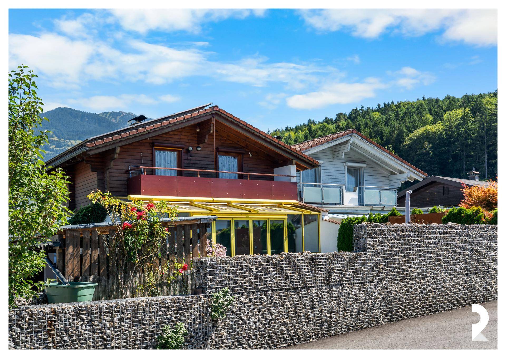 Traditionelles Holzhaus mit Balkon und angrenzendem Gebäude vor bergiger Landschaft.