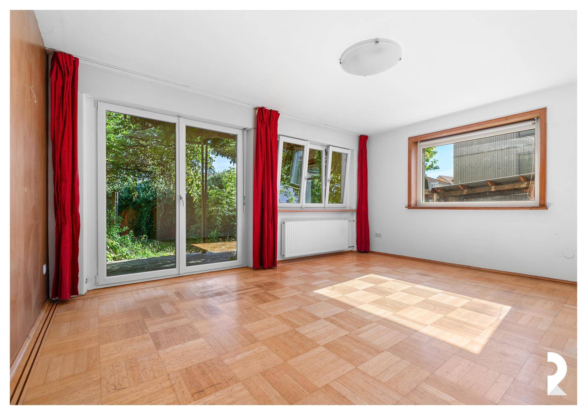 Helles Wohnzimmer mit Parkettboden, Heizkörper und großen Fenstern mit Blick ins Grüne.