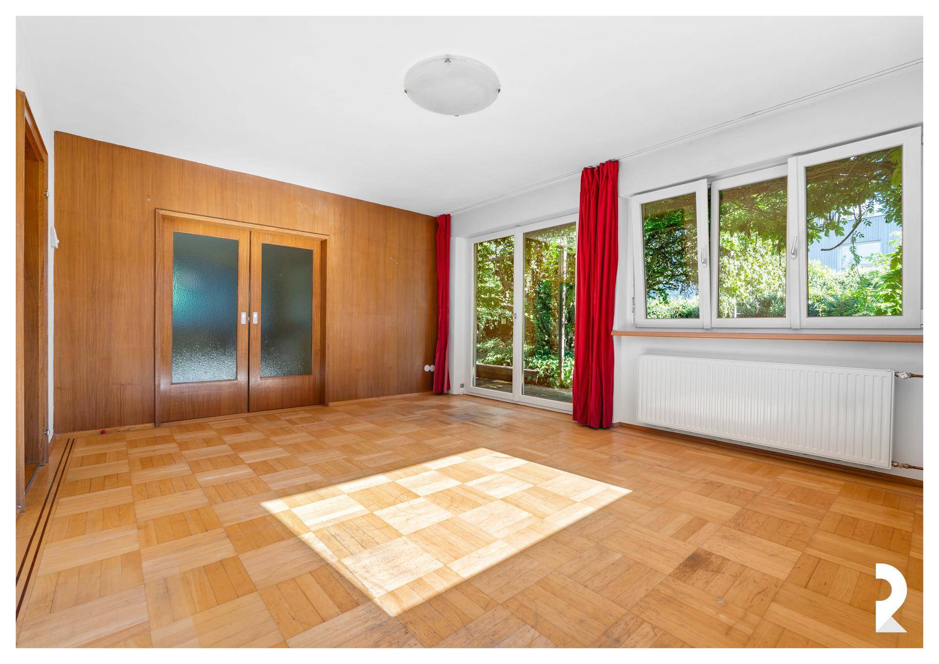 Geräumiges Wohnzimmer mit Parkettboden, Holzvertäfelung und großen Fenstern zum Garten.