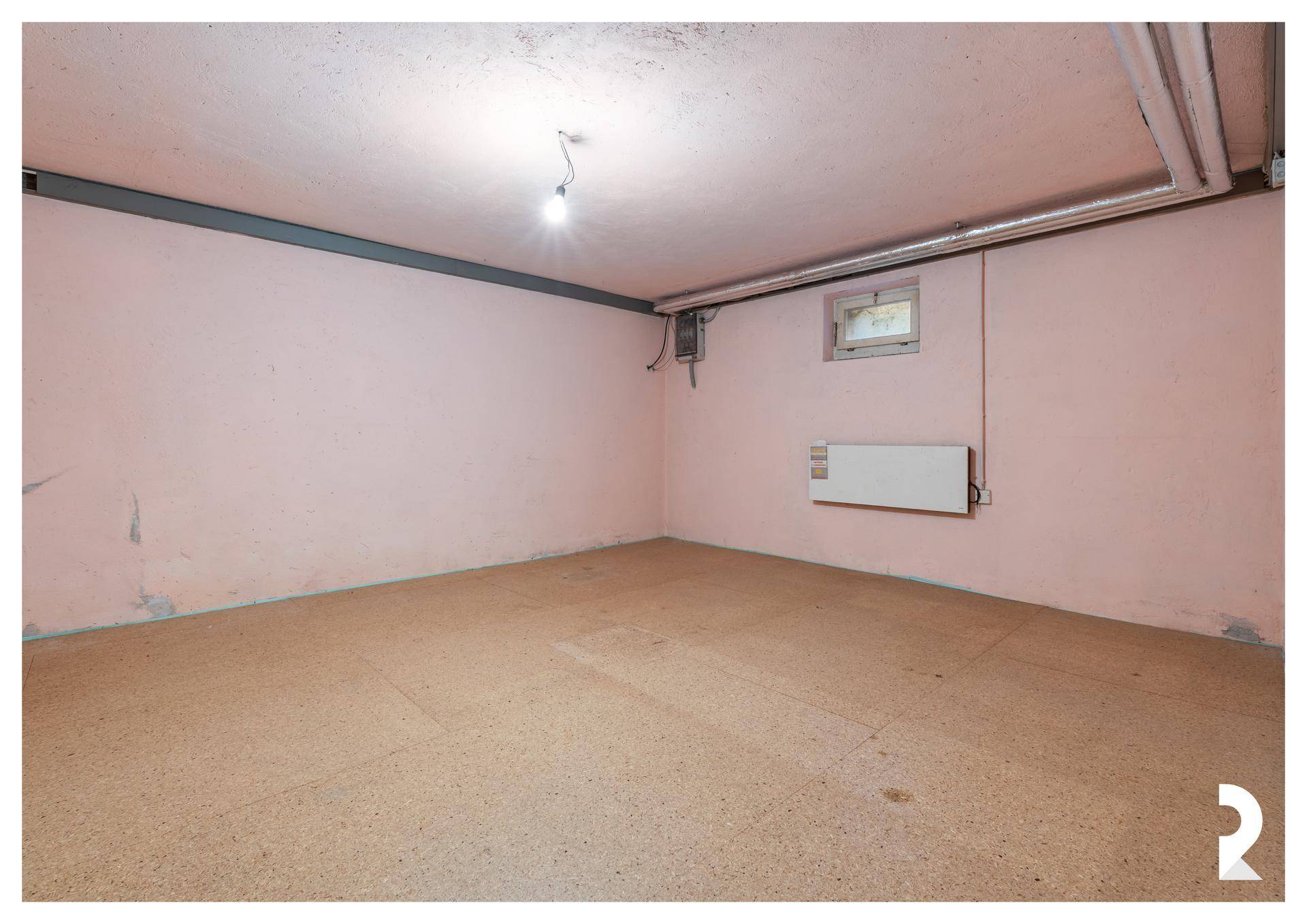 Kellerraum mit rosa gestrichenen Wänden, Betonboden, Heizkörper und sichtbaren Installationen an der Decke.