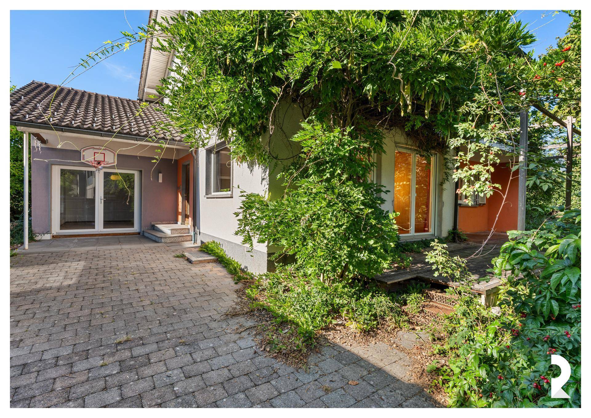 Seitenansicht des Hauses mit gepflasterter Auffahrt, viel Grün und einem Basketballkorb.