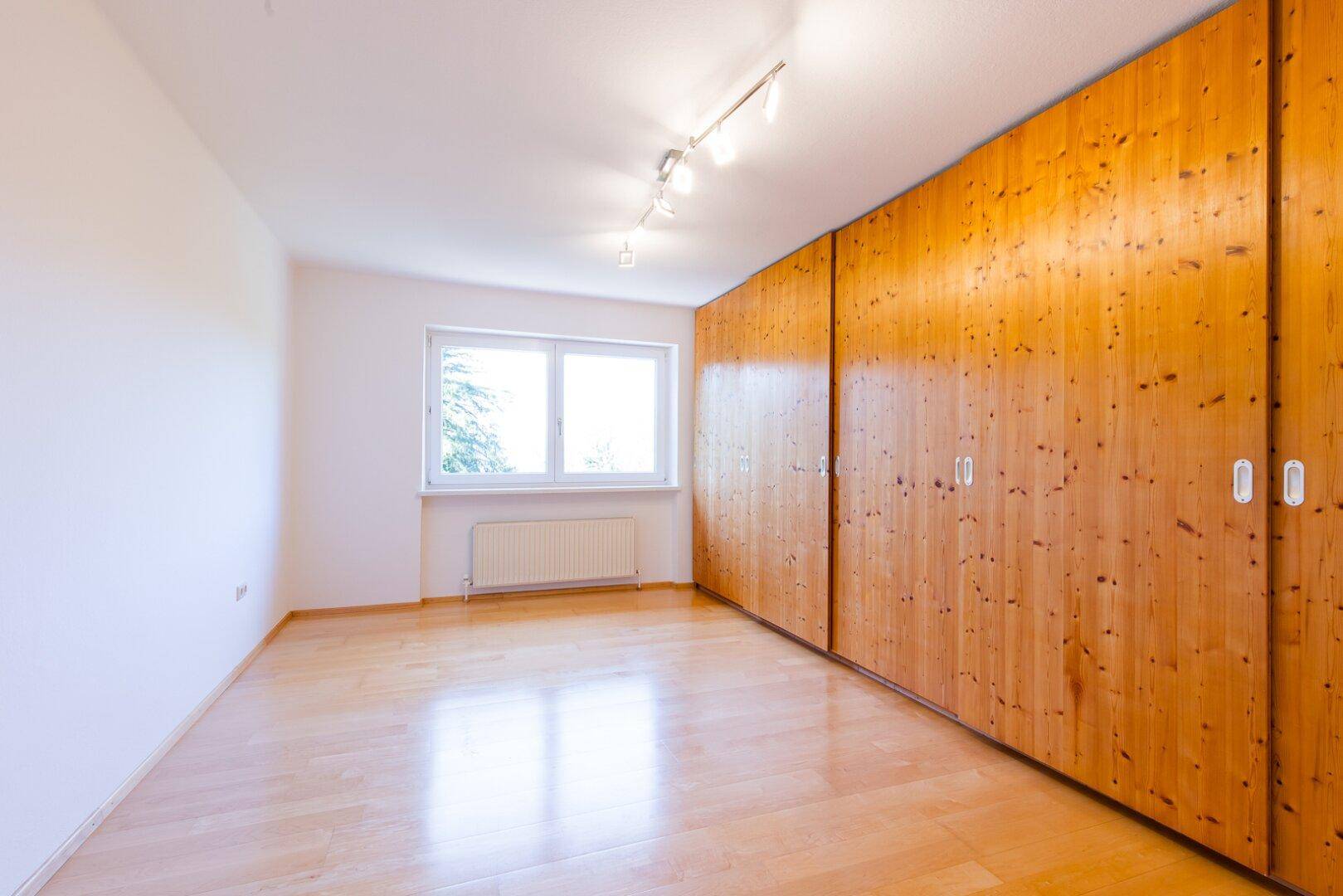 Schlafzimmer mit Holzboden, Einbauschränken und einem großen Fenster mit Blick ins Grüne.