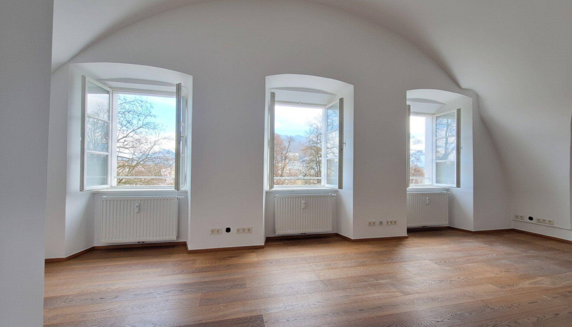 Leerer Raum mit drei großen Fenstern, Gewölbedecke und Holzboden, bietet viel Tageslicht und Weitblick.