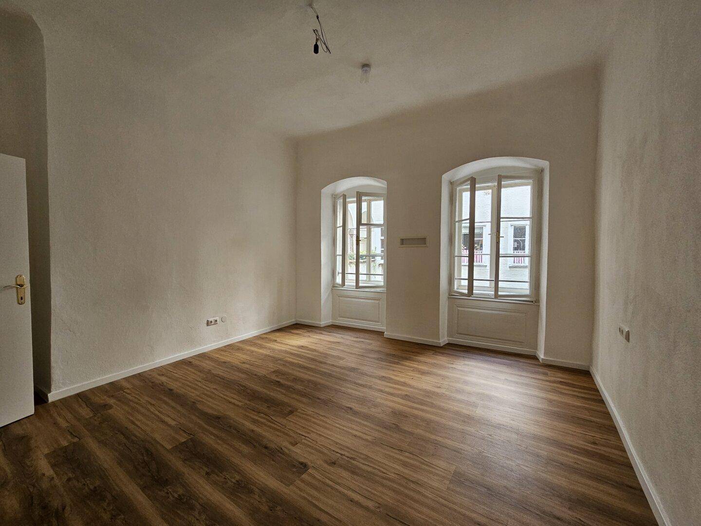 Helles Zimmer mit Holzboden und zwei hohen Fenstern, die viel Tageslicht hereinlassen.