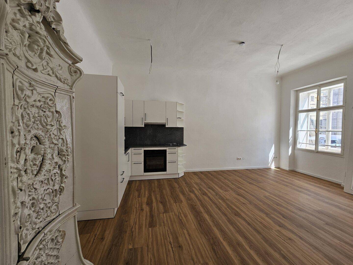 Offener Wohnbereich mit kleiner Einbauküche, Holzboden und einem traditionellen Kachelofen.