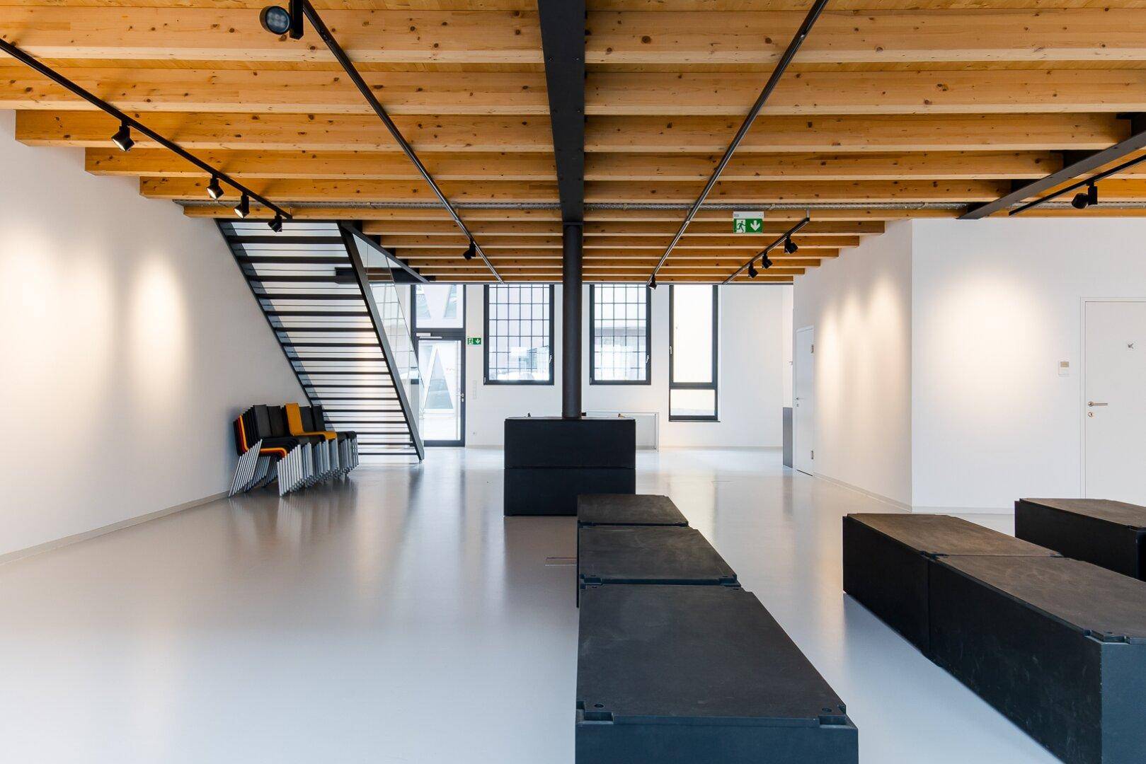 Großer, offener Raum mit hohen Decken, sichtbaren Holzbalken und einer modernen Metalltreppe.