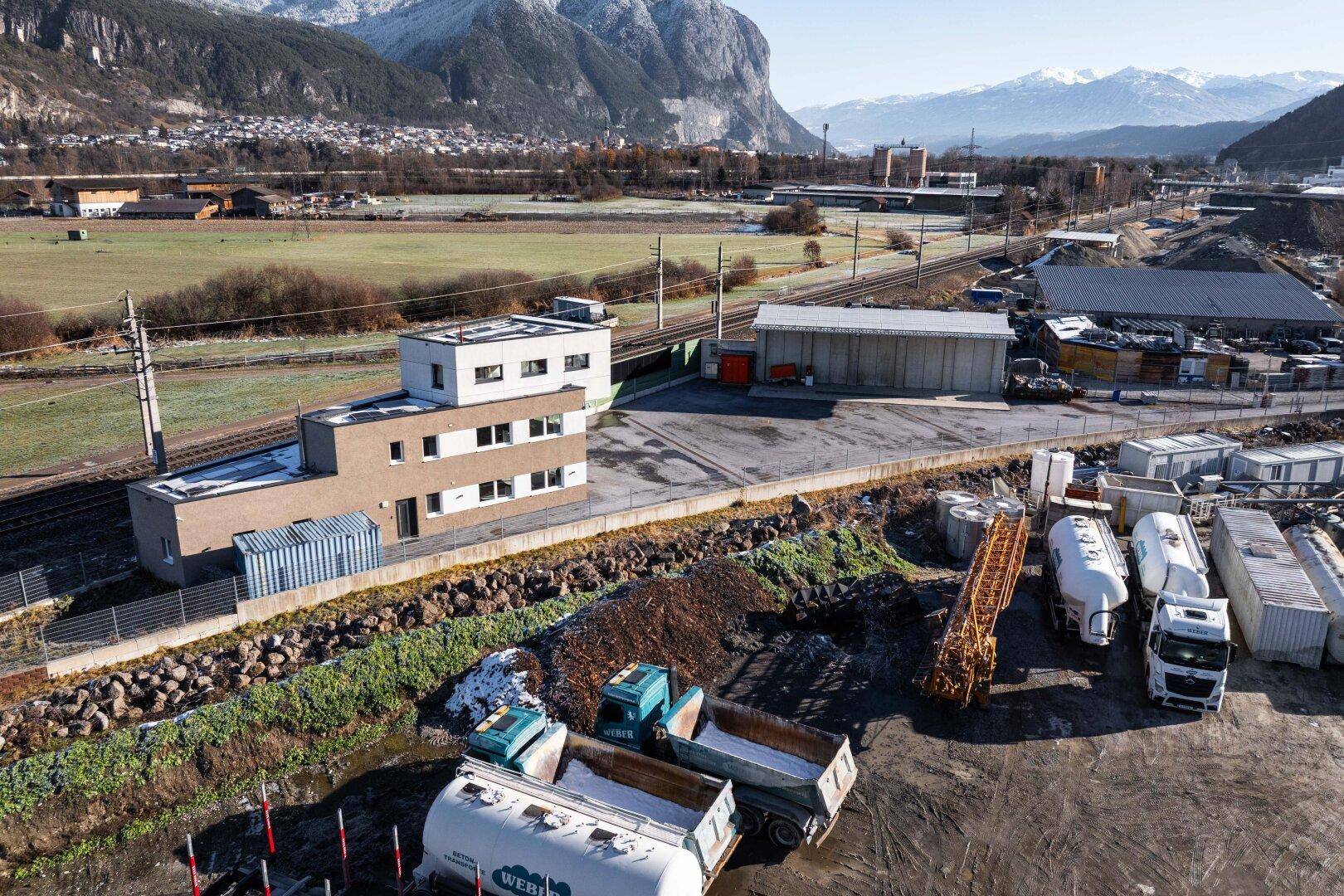 Seitenansicht des Geschäftsgebäudes mit Bahngleisen und Industrieanlagen im Hintergrund, umgeben von Bergen.