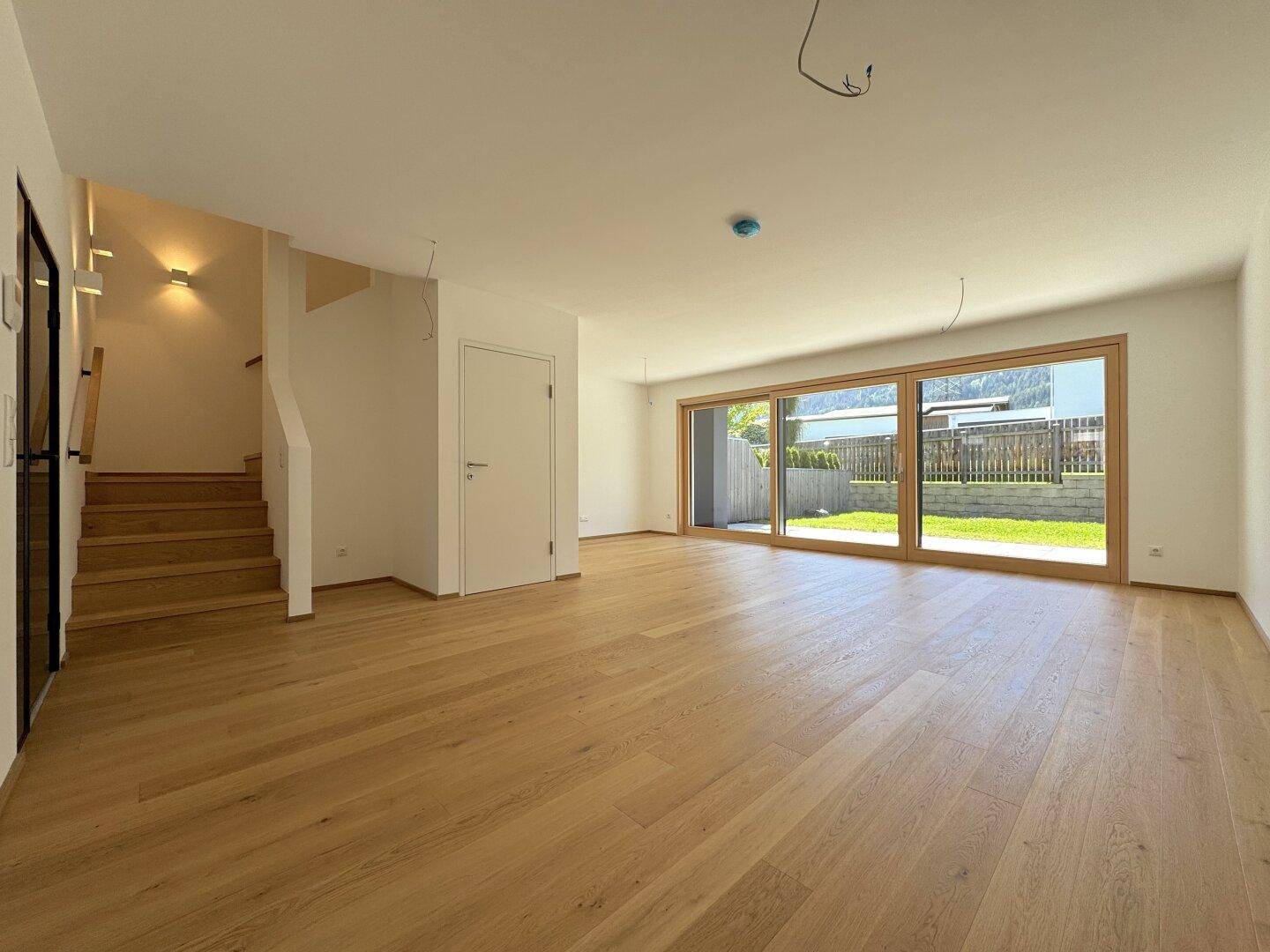 Geräumiger, unmöblierter Raum mit Holzboden, Treppe und großen Schiebetüren zum Gartenbereich.