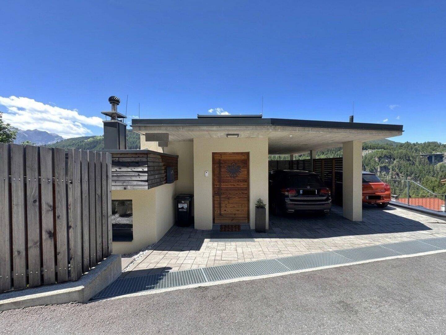 Modernes Haus mit Carport und gepflastertem Eingangsbereich in alpiner Umgebung.