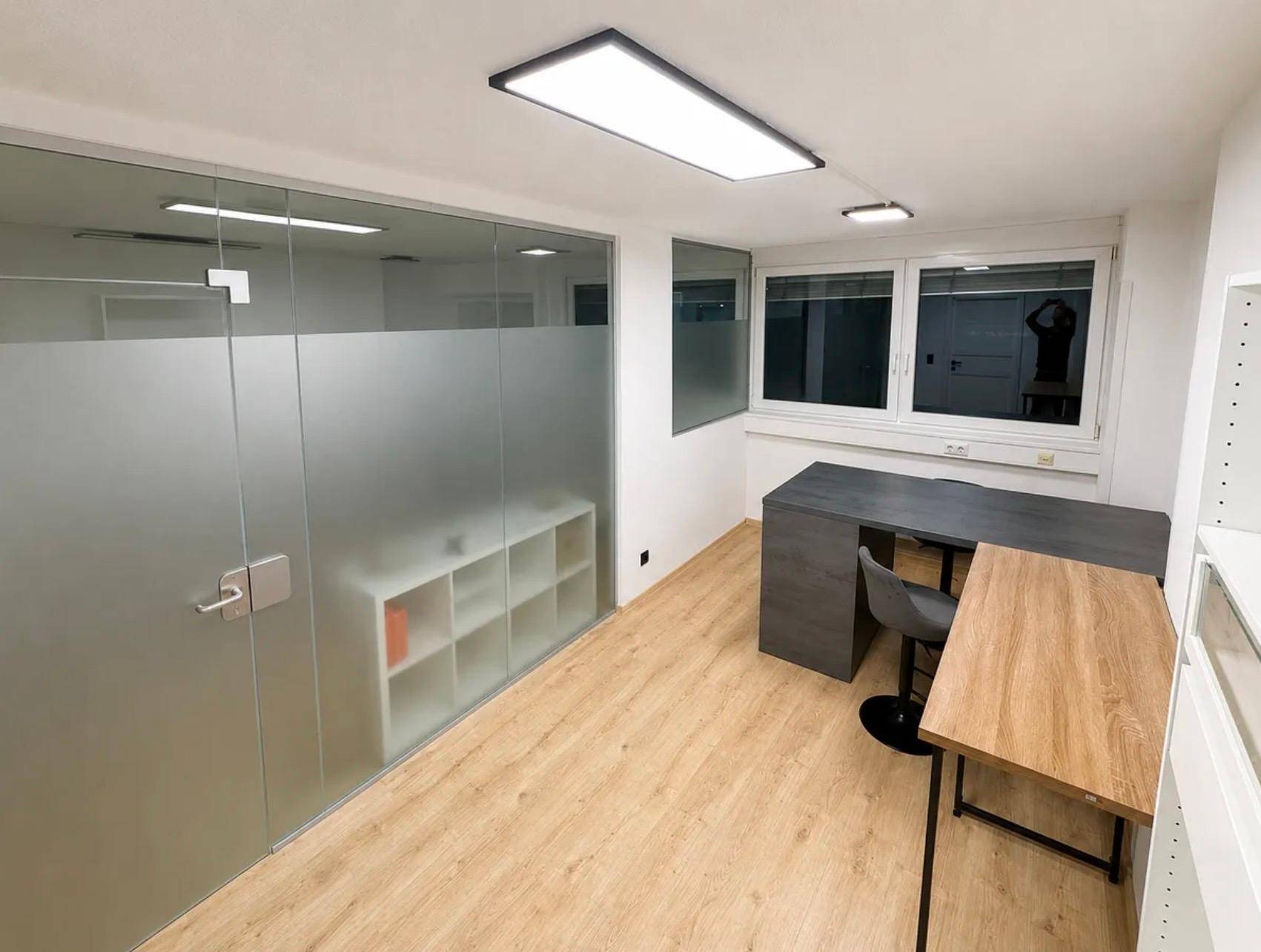 Modernes Büro mit hellem Laminatboden, großen Fenstern und einer Milchglaswand als Raumteiler.