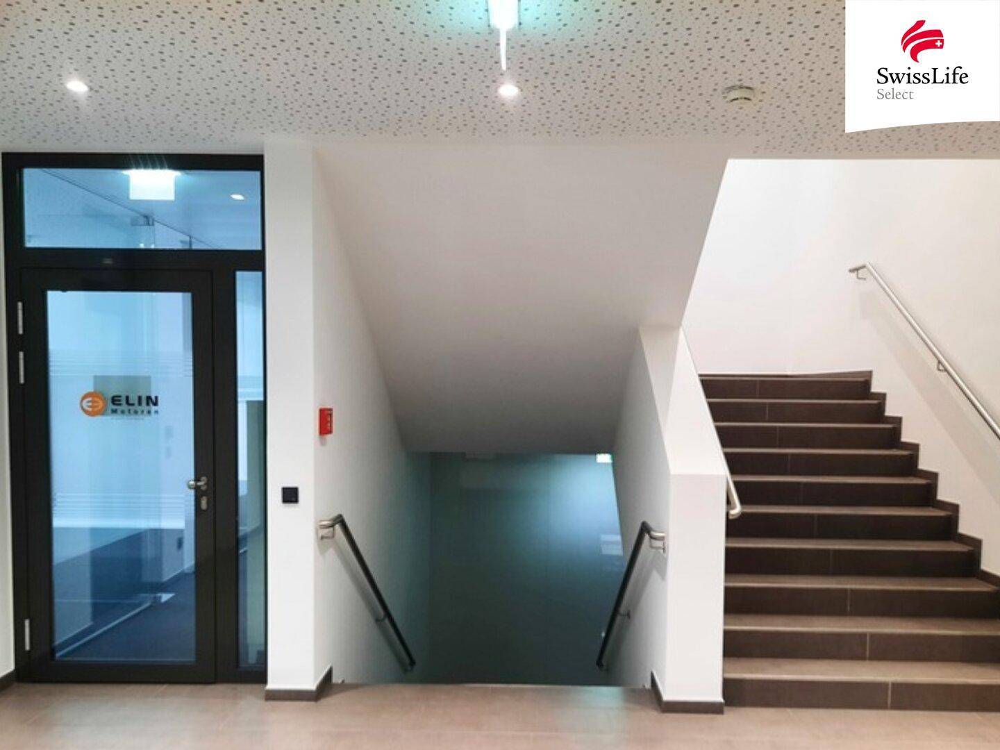 Helles Treppenhaus mit braunen Stufen, Geländer und einer Glastür zum Eingangsbereich.