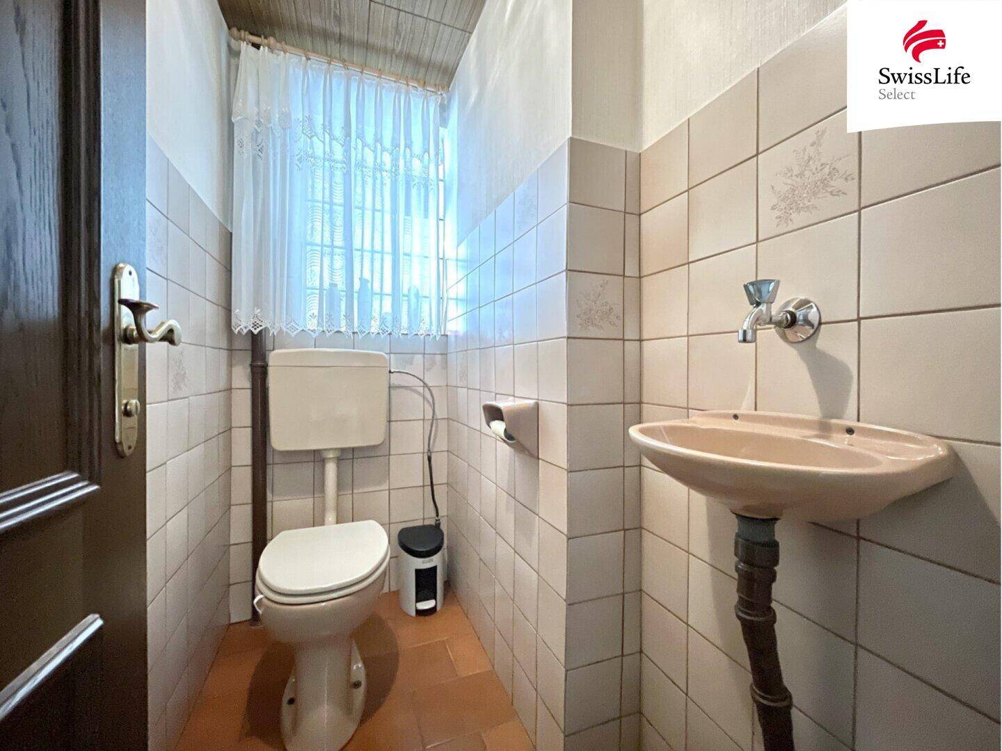 Separates WC mit Toilette, kleinem Waschbecken und traditionellen Fliesen an Wänden und Boden.