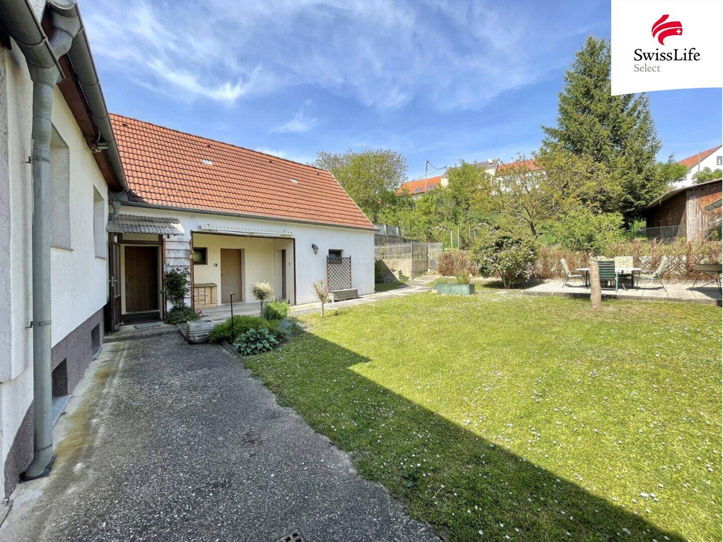Sonniger Garten mit Rasenfläche, gepflastertem Bereich und Sitzgelegenheiten im Hintergrund.