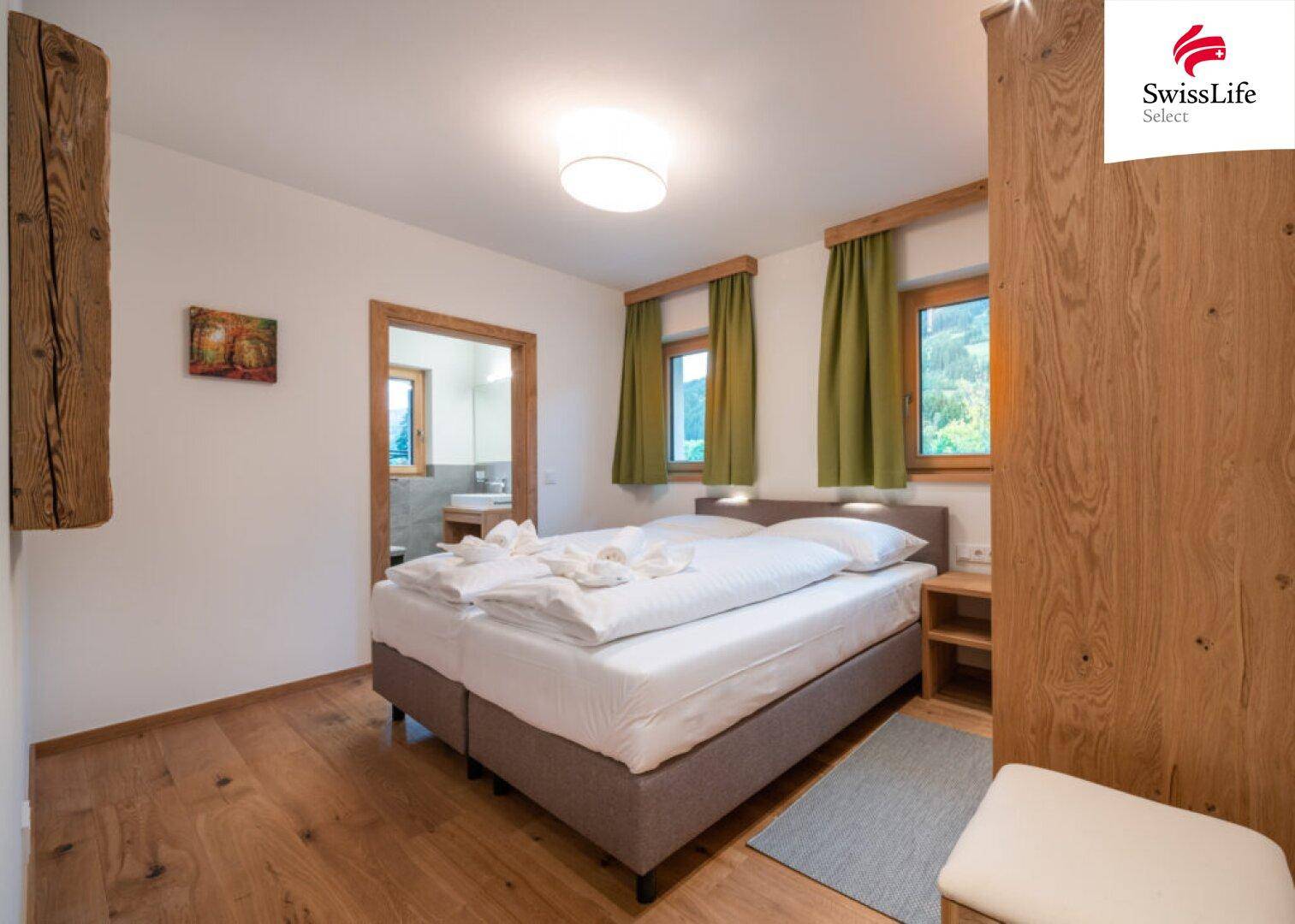 Helles Schlafzimmer mit Doppelbett, Holzboden und Zugang zu einem angrenzenden Badezimmer.