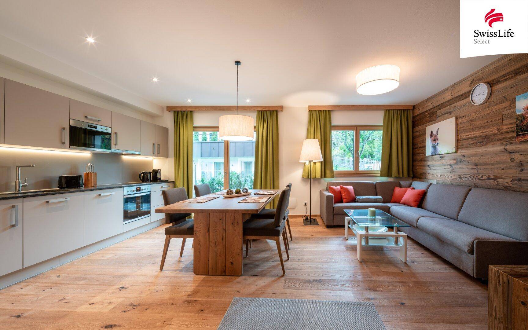 Offener Wohn-Essbereich mit moderner Küche, Holztisch und gemütlichem Sofa in alpiner Atmosphäre.