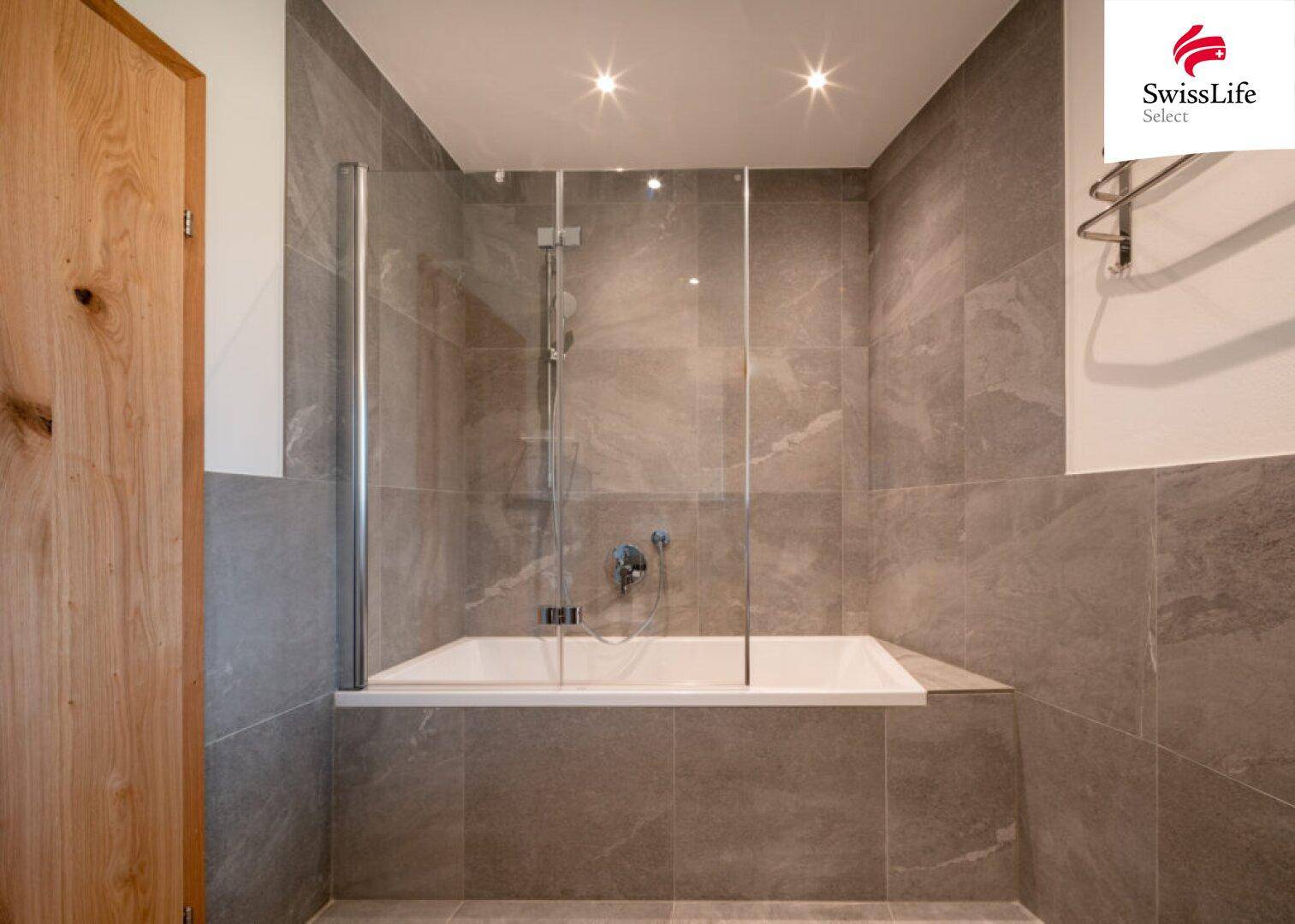 Elegantes Badezimmer mit Badewanne, Glasabtrennung und grauen großformatigen Fliesen.