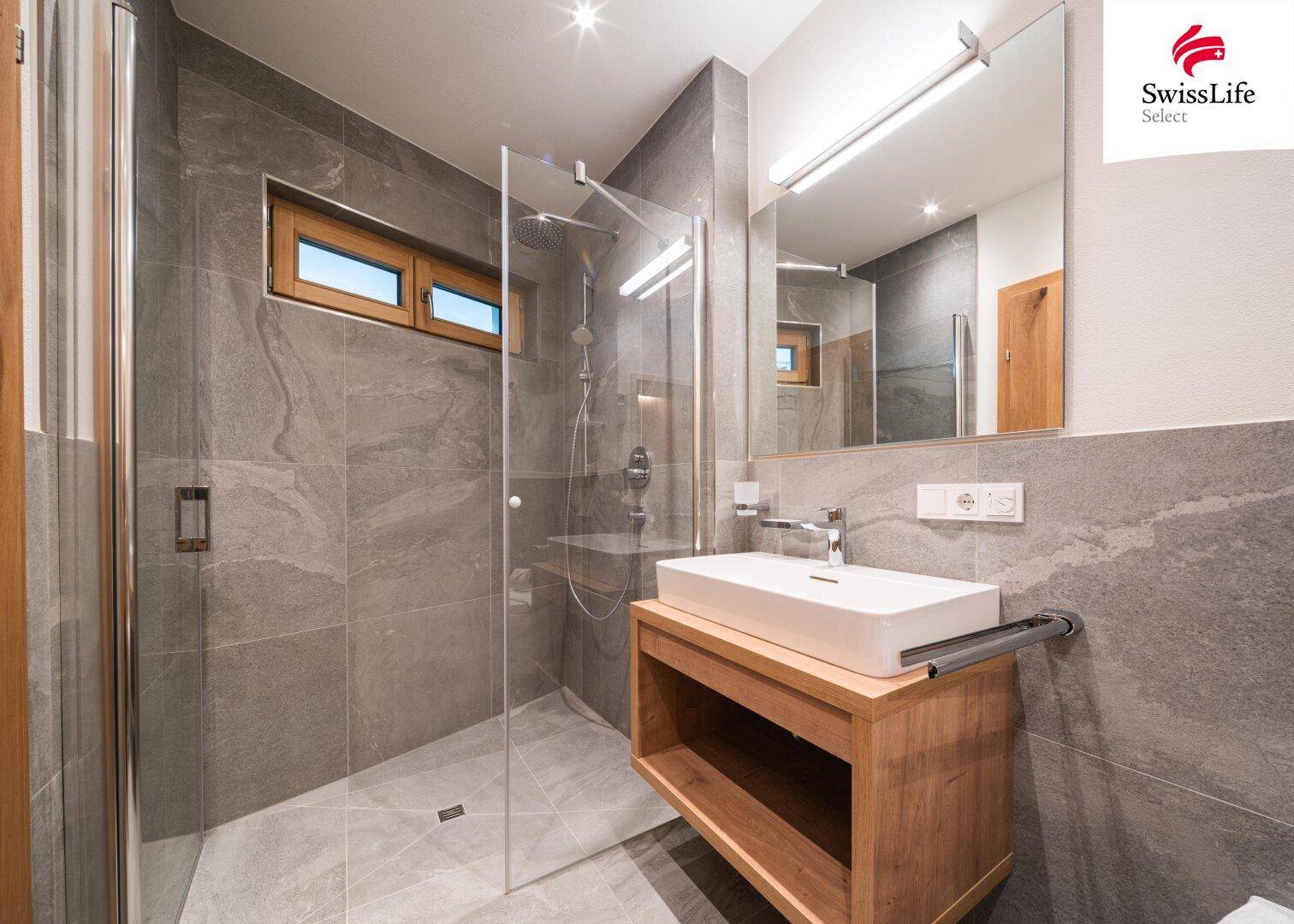 Modernes Badezimmer mit grauen Fliesen, begehbarer Dusche, Holzwaschtisch und Fenster für Tageslicht.