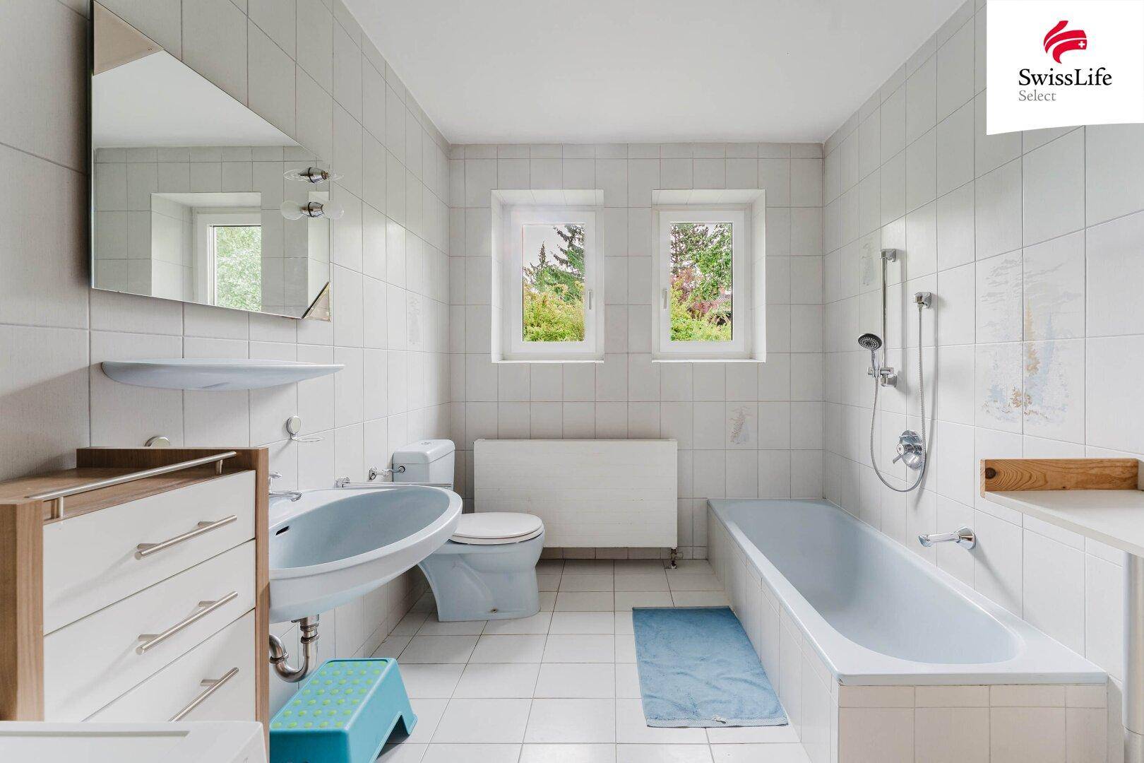 Badezimmer mit Badewanne, WC, Waschbecken und zwei Fenstern für Tageslicht.