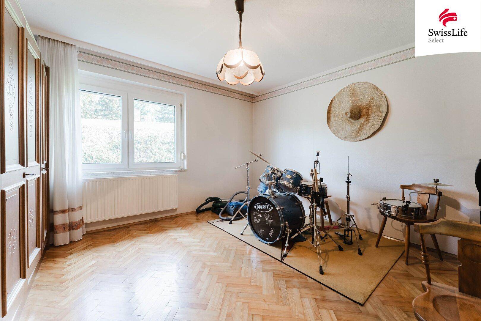 Geräumiges Zimmer mit Fischgrätparkett, großem Fenster und Schlagzeug-Set.