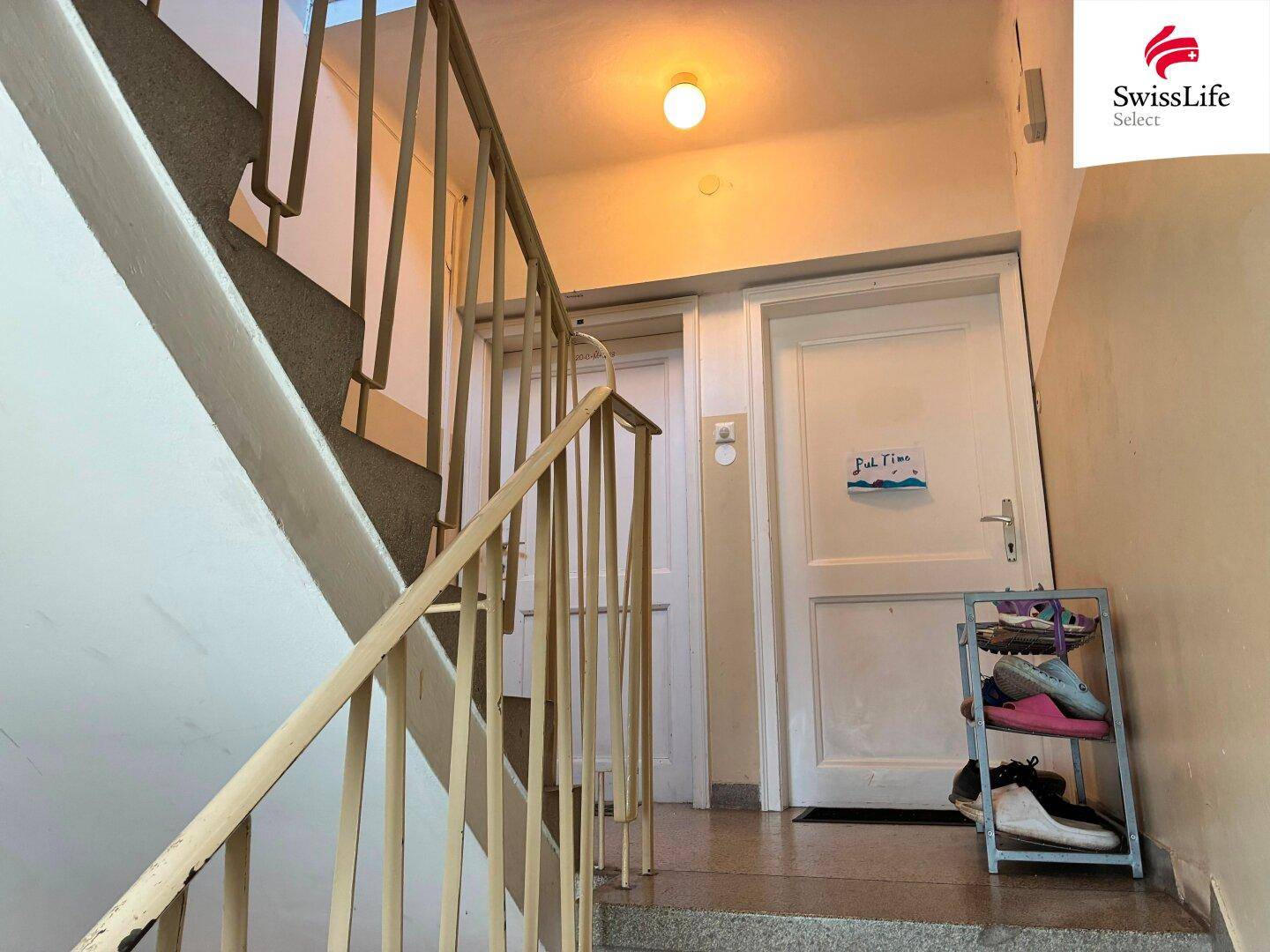 Treppenhaus mit Steinstufen, Geländer und zwei Wohnungstüren, eine davon mit Schuhregal.