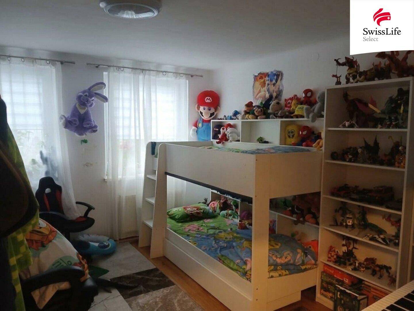 Kinderzimmer mit Etagenbett, vielen Spielsachen und zwei Fenstern, die für natürliches Licht sorgen.