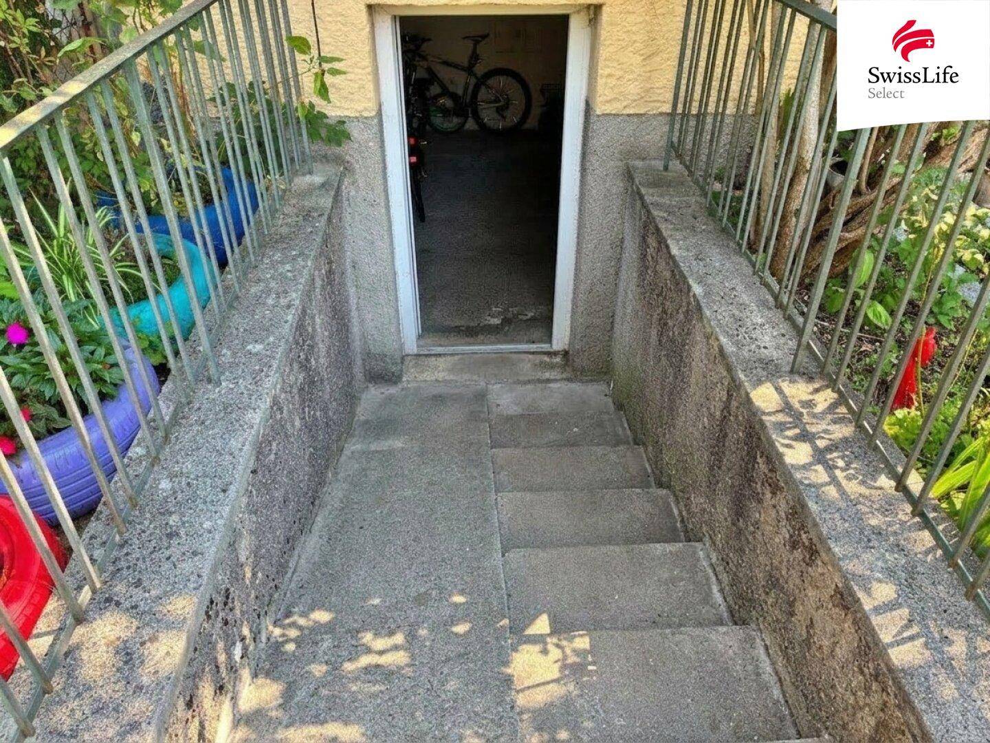 Außentreppe aus Beton, die zum Kellereingang des Gebäudes führt, umgeben von Pflanzen.