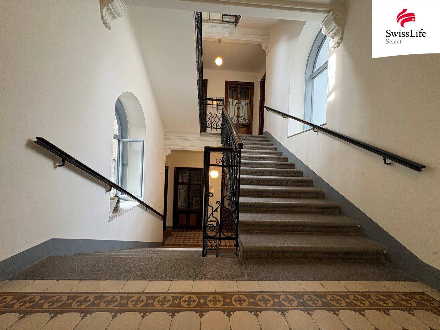Historisches Treppenhaus mit Steinstufen, schmiedeeisernem Geländer und großen Fenstern.