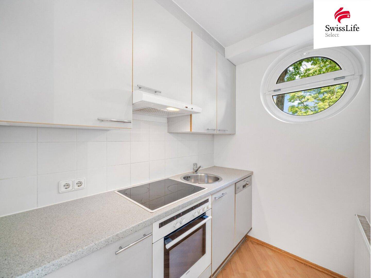 Kompakte Küche mit weißen Schränken, Einbaugeräten und einem charakteristischen runden Fenster.
