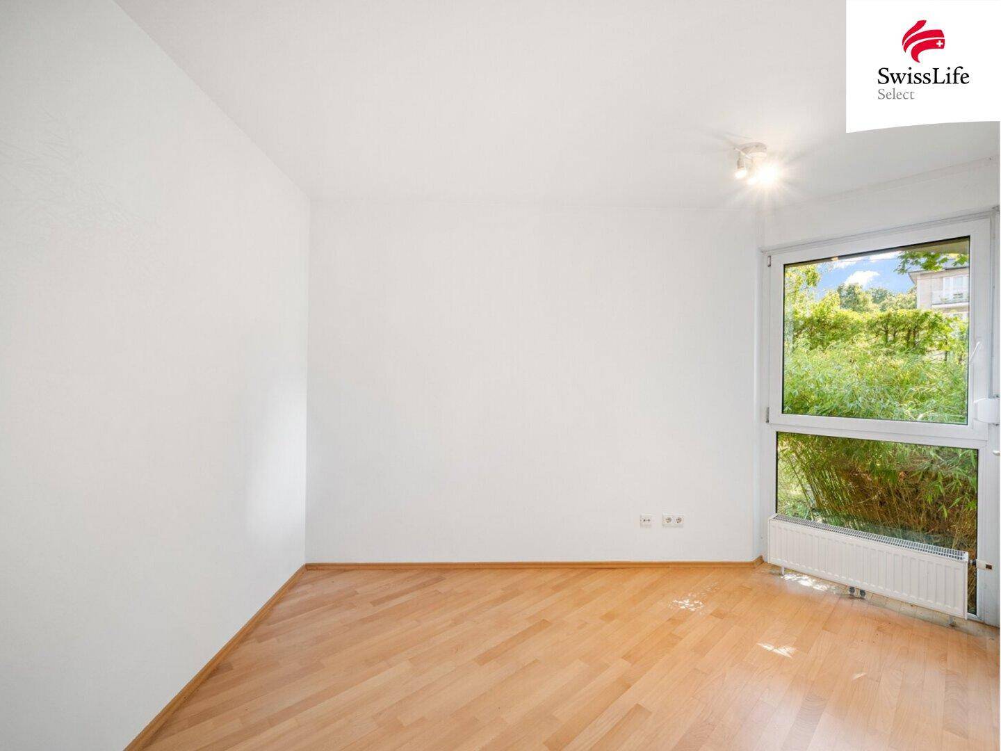 Leeres Zimmer mit Holzfußboden und großem Fenster, das viel Tageslicht hereinlässt.