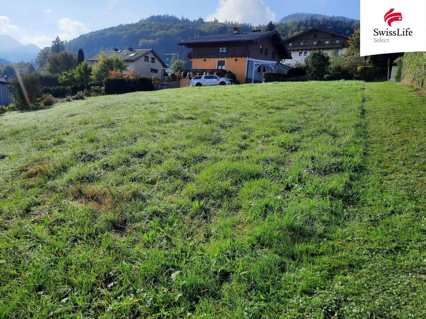 Sonnenbeschienenes grünes Grundstück mit Blick auf umliegende Häuser und Berge.