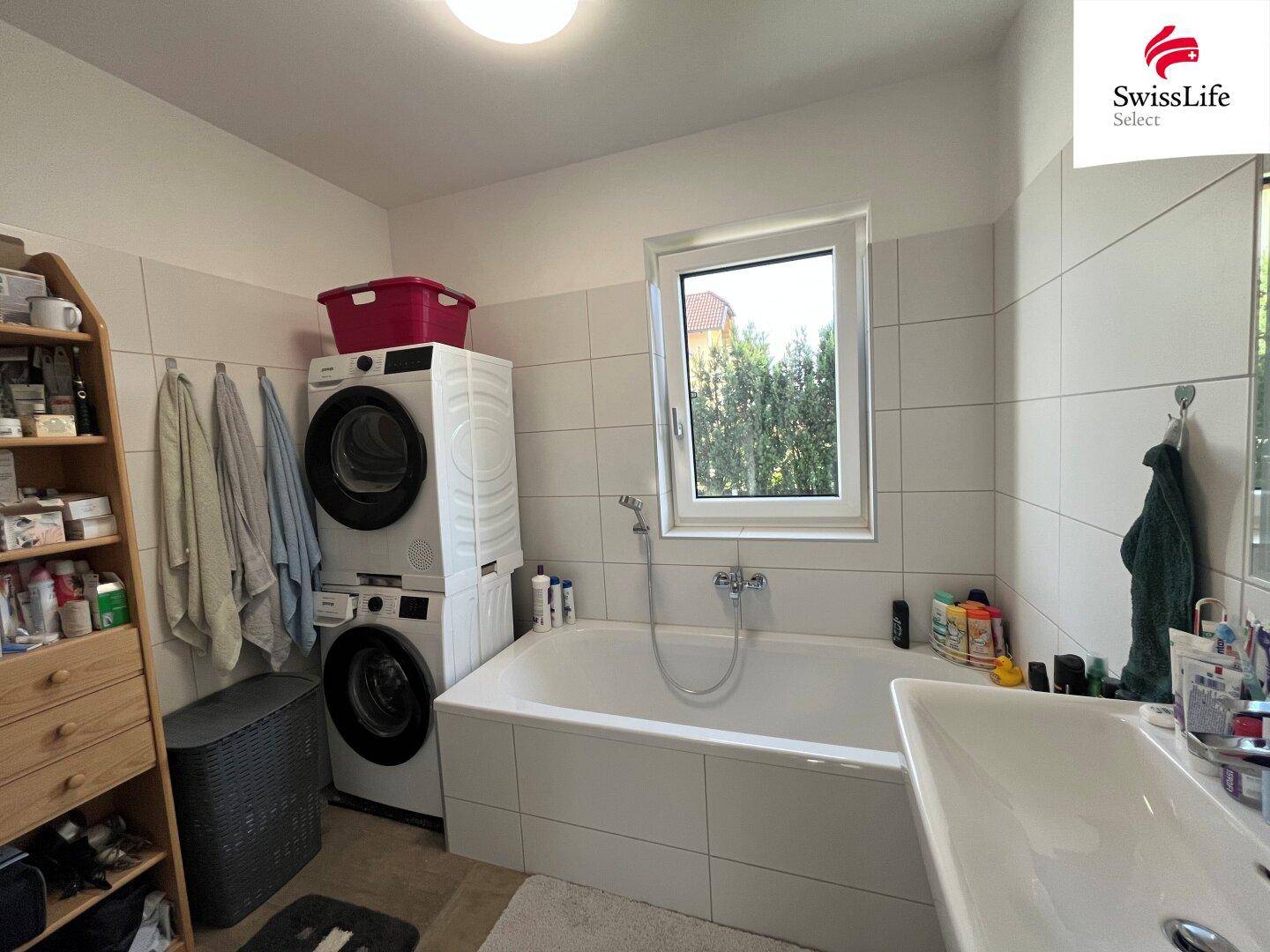 Badezimmer mit Badewanne, Waschmaschine und Trockner sowie einem Fenster.