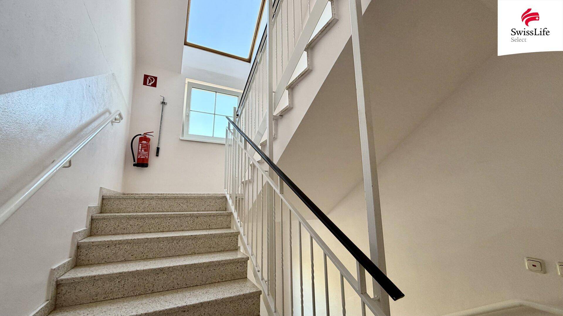 Treppenhaus mit Terrazzostufen, Geländer und Fenster, das Tageslicht hereinlässt.