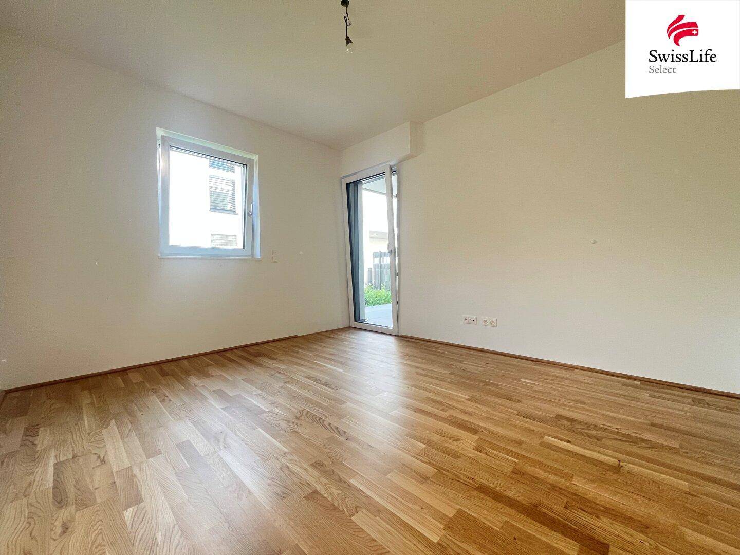 Leeres Schlafzimmer mit hellem Holzparkettboden und Fenster mit Zugang nach draußen.