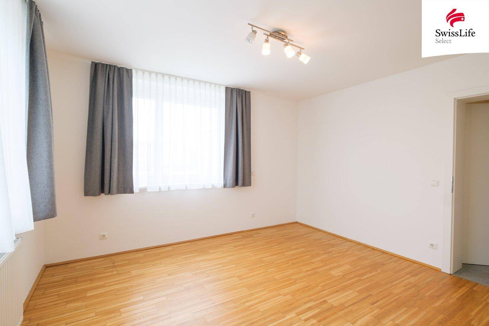 Helles Zimmer mit Parkettboden, weißen Wänden und einem großen Fenster mit Vorhängen.