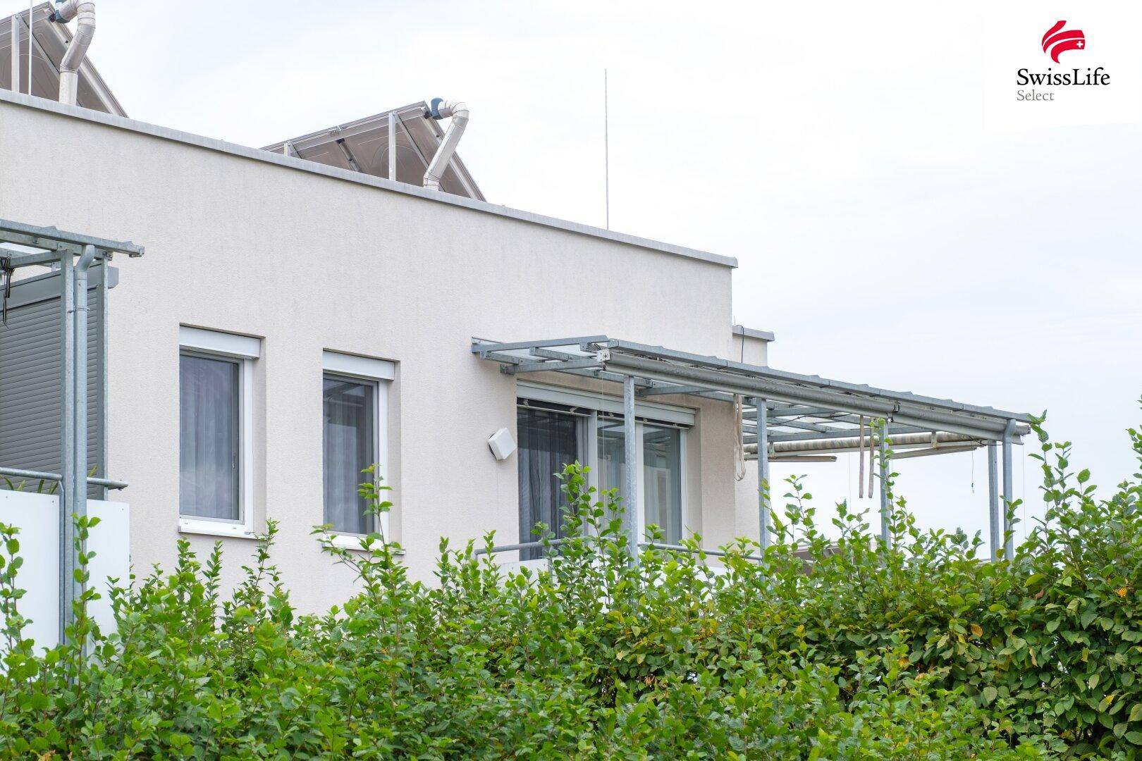 Gebäudefassade mit Balkon, Jalousien und sichtbaren Photovoltaik-Modulen auf dem Dach.