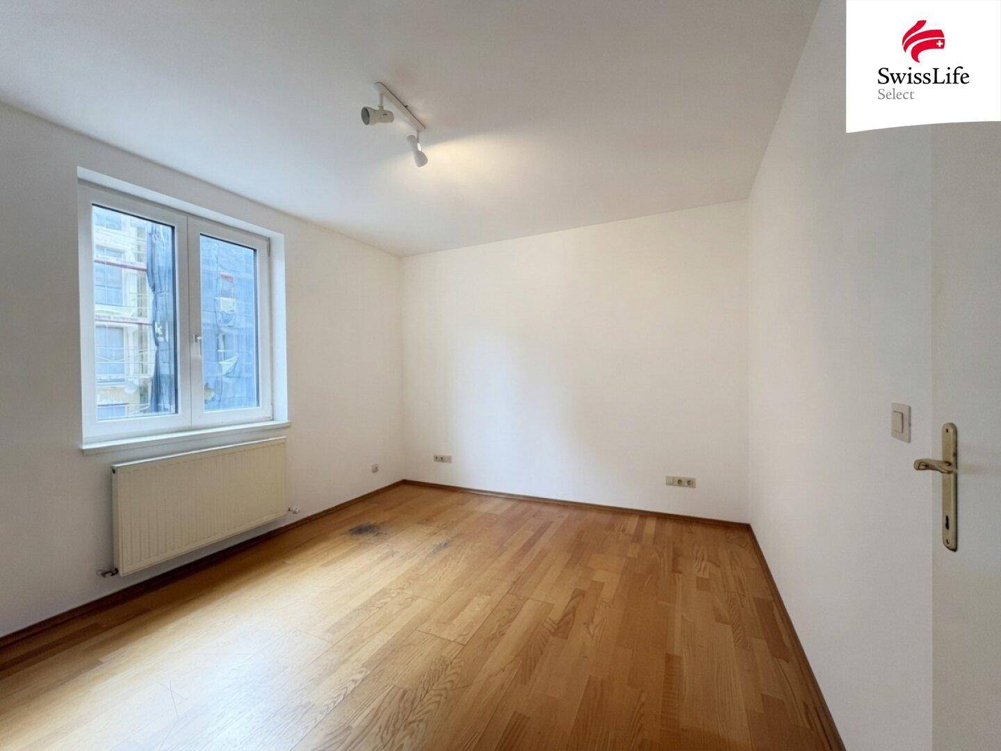 Helles Schlafzimmer mit Parkettboden und einem großen Fenster, das viel Licht hereinlässt.