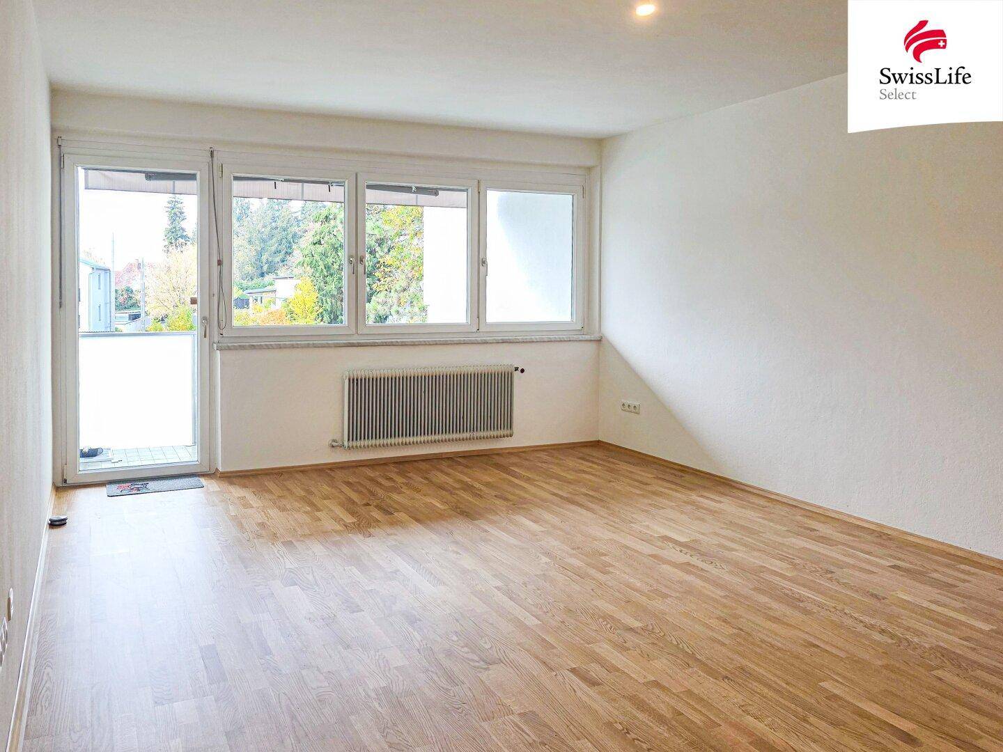 Helles Wohnzimmer mit Parkettboden, großem Fenster und Zugang zu einem Balkon.