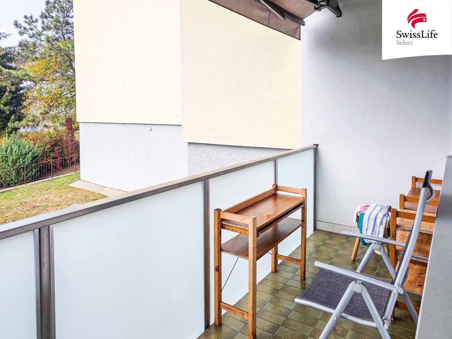 Balkon mit Fliesenboden, Glasbrüstung und einfacher Bestuhlung, umgeben von Grün.