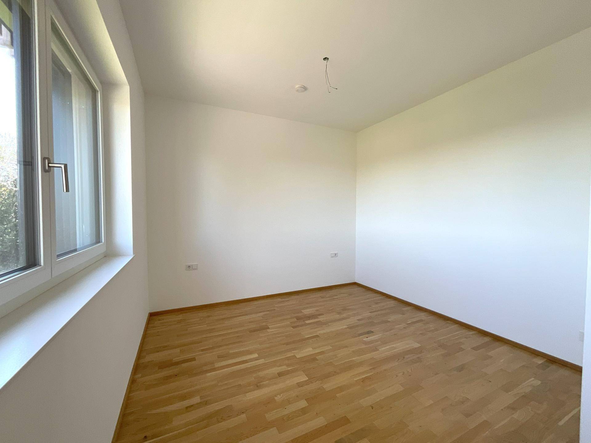 Leerer Raum mit Parkettboden und einem Fenster, das den Blick ins Grüne freigibt.