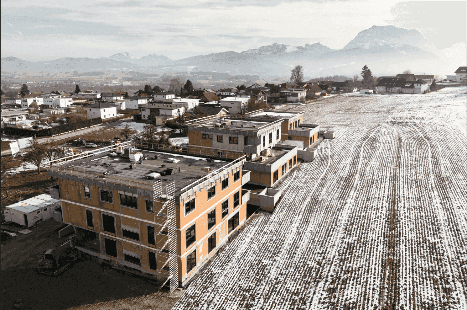 Luftaufnahme einer Neubauanlage im Winter mit verschneiten Feldern und Bergpanorama im Hintergrund.
