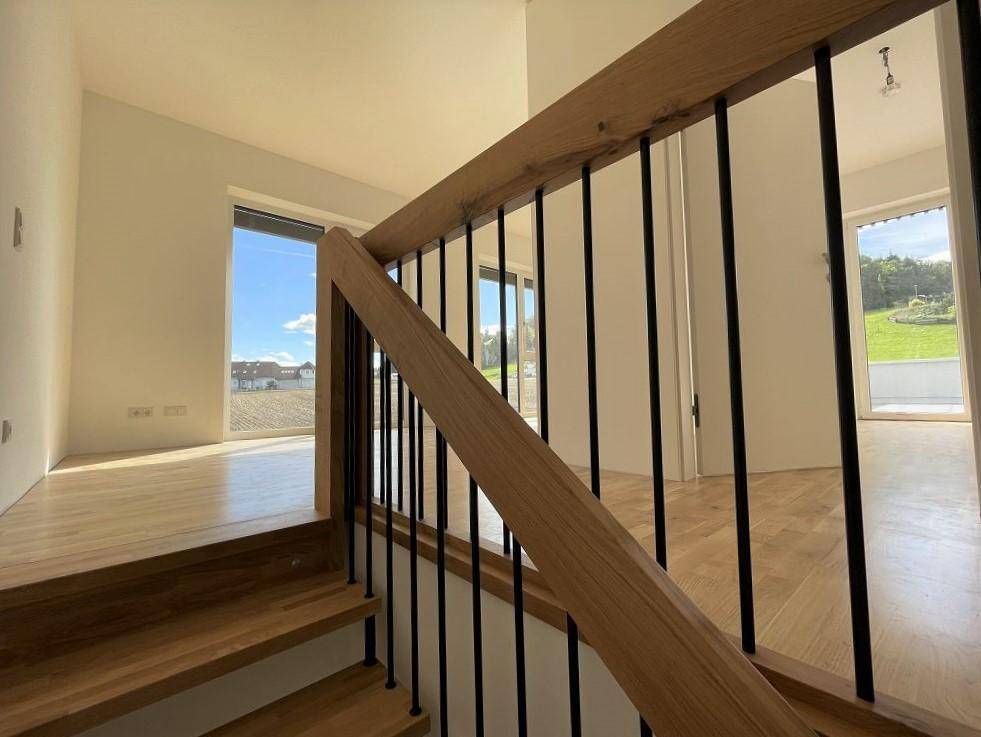 Blick von der Holztreppe in einen hellen Raum mit großen Fenstern und grünem Ausblick.