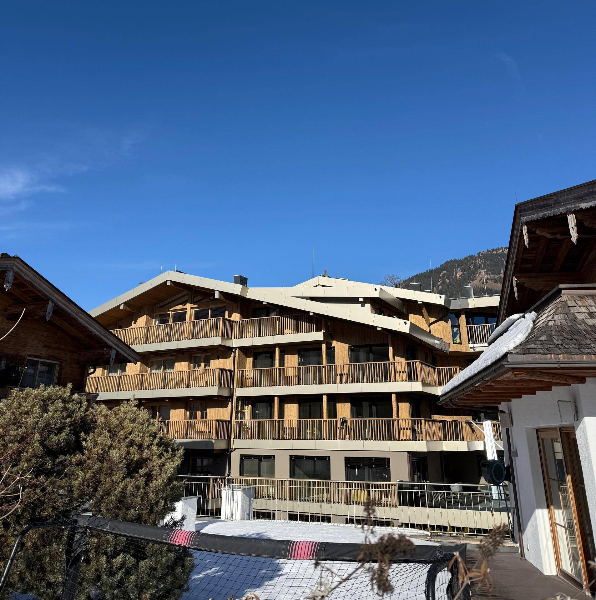 Modernes Gebäude mit Holzfassade und Balkonen, umgeben von Schnee und traditionellen Häusern.